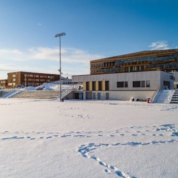 Klubbhus ved snødekt fotballbane. Skolebygg i bakgrunnen