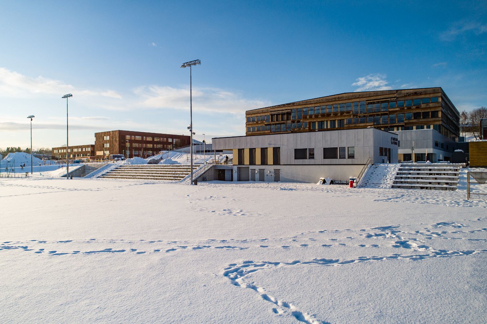 Klubbhus ved snødekt fotballbane. Skolebygg i bakgrunnen